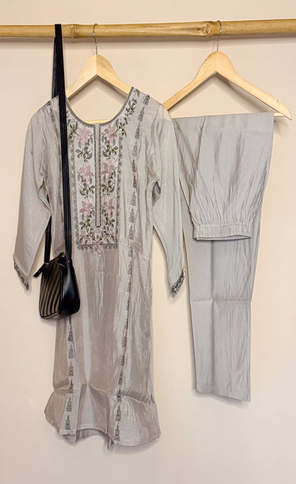 Gray embroidered top and pants with a black bag on hangers against a beige wall.